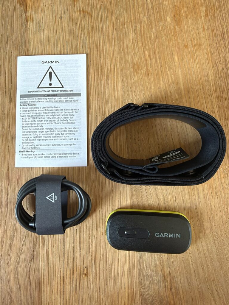 Garmin HRM 600 mit Sicherheitshinweisen, Gurt, aufgerolltem Ladekabel und Modul auf einem Holztisch