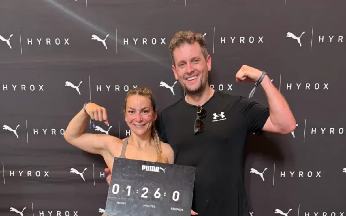 Alex und Geri vor der HYROX-Fotowand in Berlin, Geri hält eine Tafel mit ihrer Zielzeit