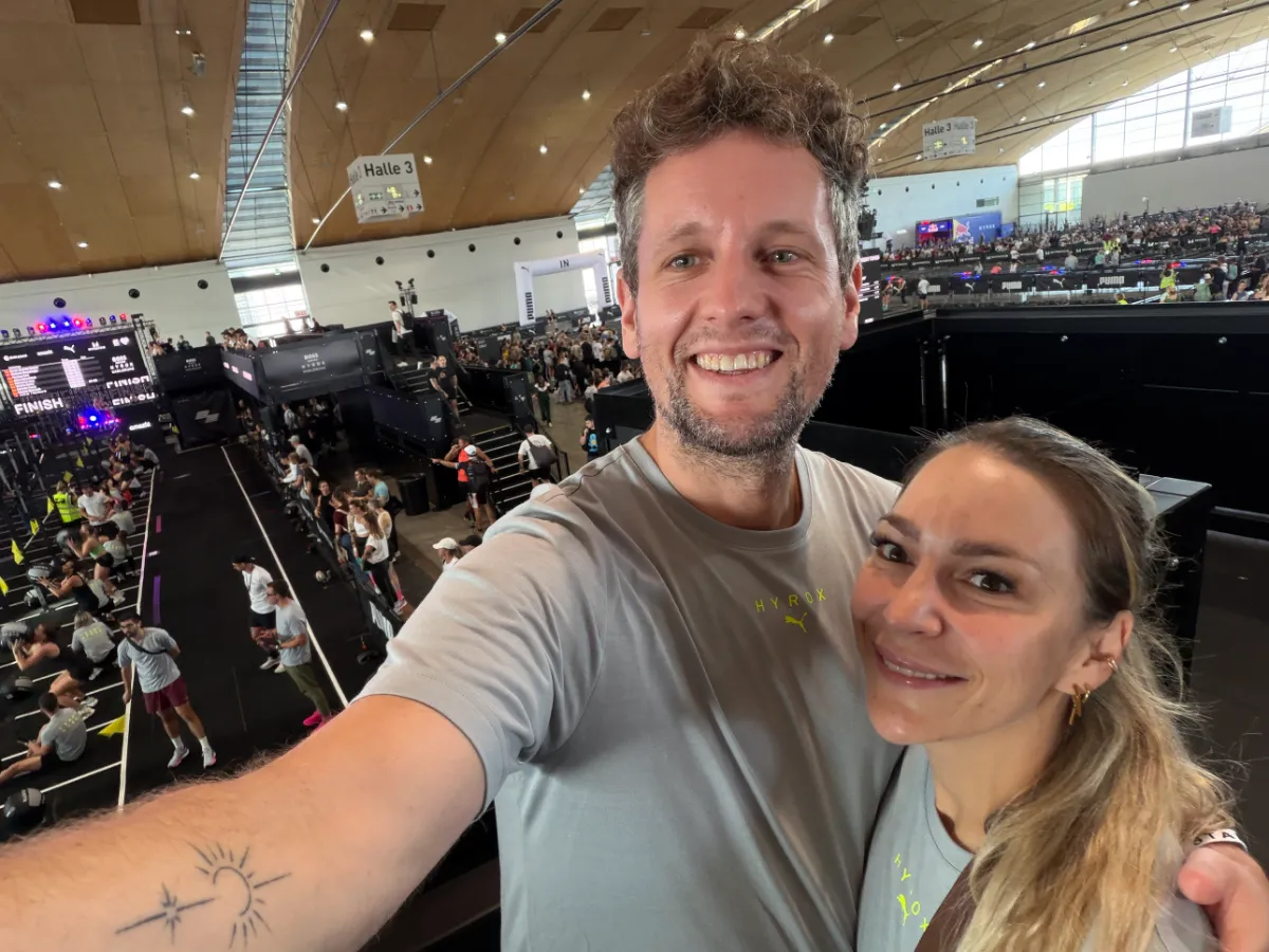 Alex und Geri machen ein Selfie auf der Tribüne über Halle 3 bei HYROX Karlsruhe 2026, unten das Rennfeld